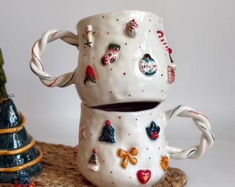 May include: Two white ceramic mugs stacked on top of each other, decorated with Christmas-themed embellishments. Each mug has a twisted handle with red and green accents. The mugs are adorned with miniature ornaments.