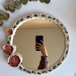 May include: Circular decorative mirror with a white ceramic frame. The frame features green leaves and red flowers. The mirror reflects a person holding a phone. Eucalyptus leaves are in the background.