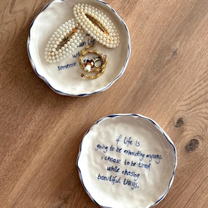 May include: Two white ceramic trinket dishes with blue trim and handwritten text. One dish holds pearl hair clips and gold jewelry. The other dish has the quote: "if life is going to be exhausting anyway, I choose to be tired while chasing beautiful things."