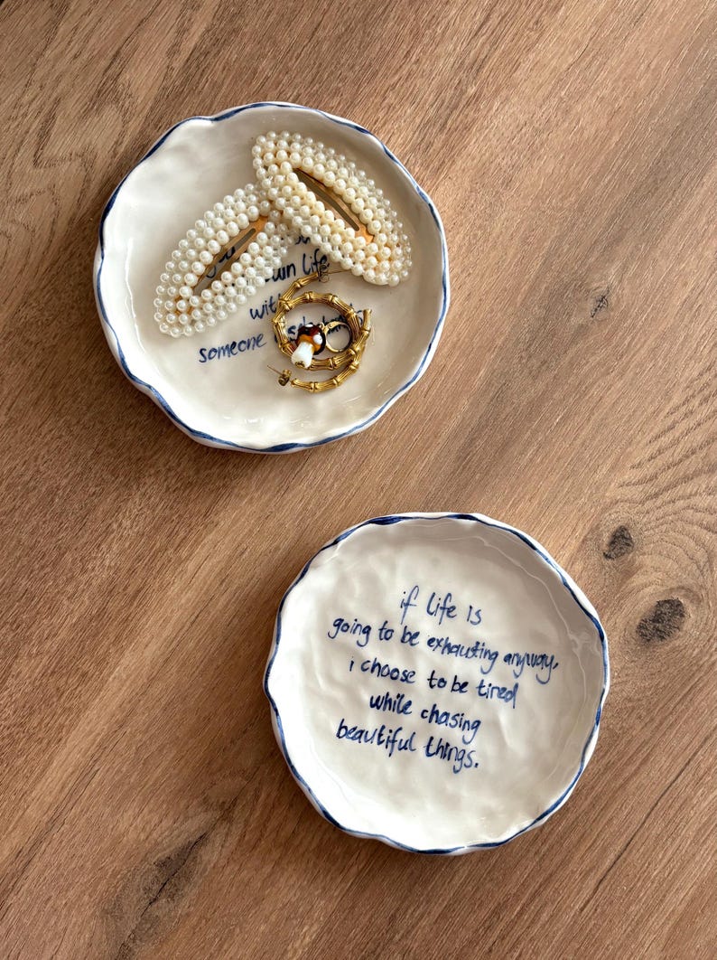 May include: Two white ceramic trinket dishes with blue trim and handwritten text. One dish holds pearl hair clips and gold jewelry. The other dish has the quote: "if life is going to be exhausting anyway, I choose to be tired while chasing beautiful things."