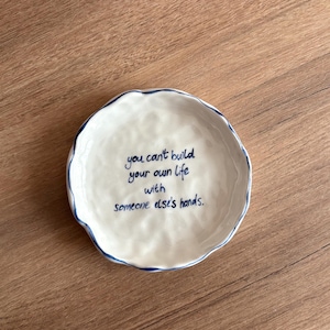 Könnte beinhalten: Eine weiße Keramikschale mit gewelltem Rand und blauem Rand. Die Schale zeigt das handgeschriebene Zitat: „you can't build your own life with someone else's hands.“ Die Schale steht auf einer Holzoberfläche.