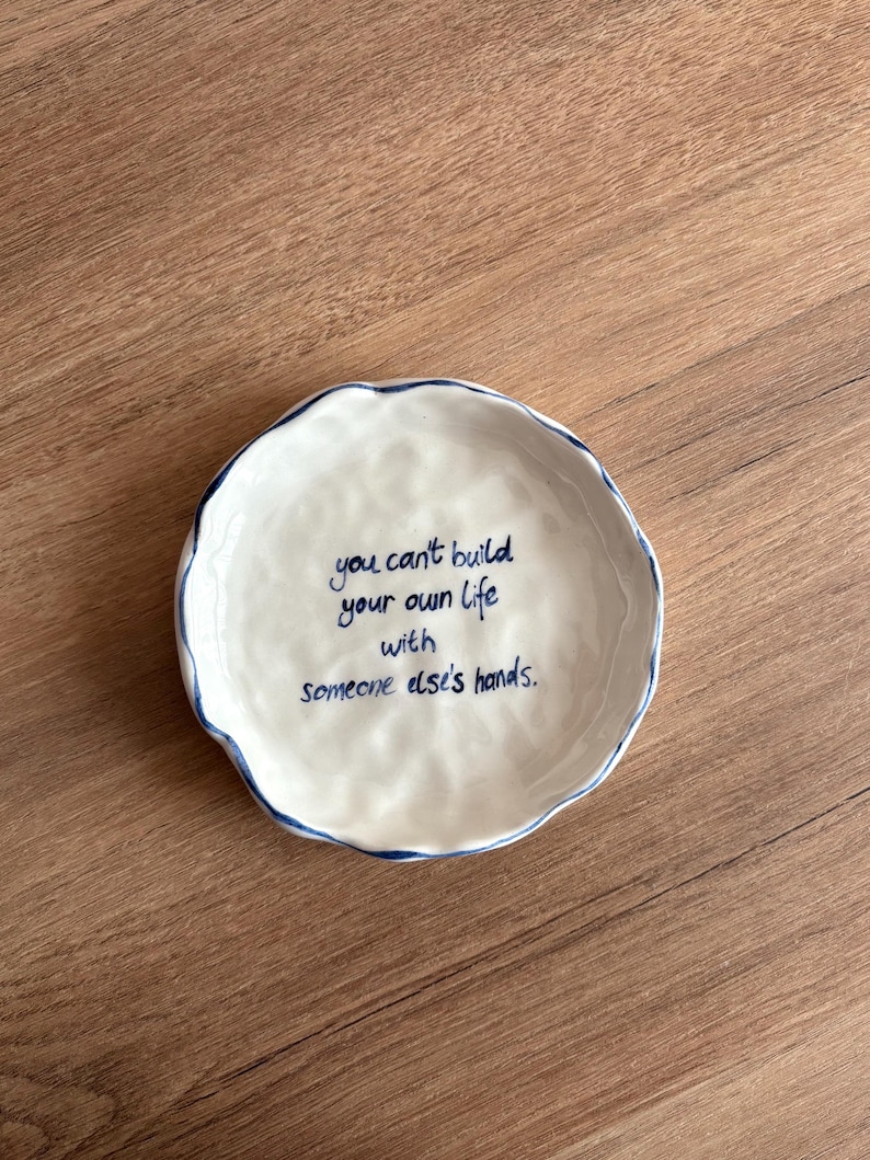 May include: A white ceramic dish with a wavy rim and a blue border. The dish features the handwritten quote, "you can't build your own life with someone else's hands." The dish is on a wooden surface.