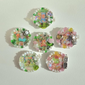 May include: Six small round resin trinket dishes with white pearl borders. Each dish features a different colorful image of cartoon characters and a variety of embellishments, including pearls, glitter, and charms.