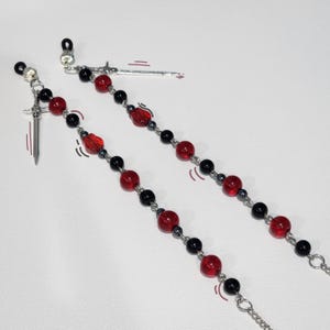 May include: Two eyeglass chains with red and black beads, and silver sword charms. The chains have a mix of round and faceted red beads, interspersed with black beads. The chains are connected with silver chain links.