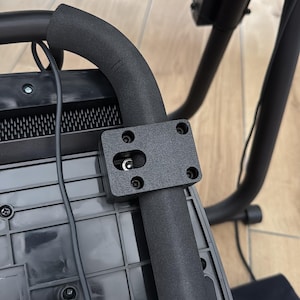 May include: Close-up of a black device with a curved, padded handle and a rectangular mounting plate. The plate is attached with screws and holds a small, silver component. Wires and a black box are also visible.