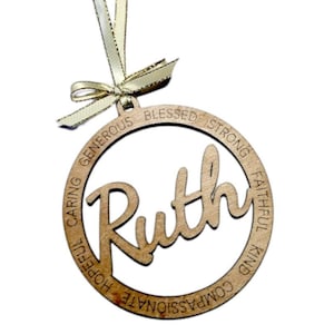 Puede incluir: Un adorno de madera con el nombre "Ruth" en una fuente cursiva dentro de un círculo. El círculo está rodeado por las palabras "Bendecido, Fuerte, Fiel, Amable, Compasivo, Apasionado, Esperanzador, Cuidadoso, Generoso".