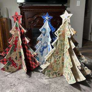 May include: Three fabric Christmas tree decorations standing upright. The left tree features a red truck and buffalo plaid pattern. The center tree displays blue ornaments on a white background. The right tree shows a winter forest scene with snowflakes.