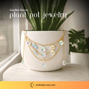 May include: A white ceramic plant pot adorned with gold-colored chains and iridescent crystal beads. The text "Garden Decor plant pot jewelry" is displayed above the pot. The jewelry hangs from the rim of the pot, adding a decorative touch.