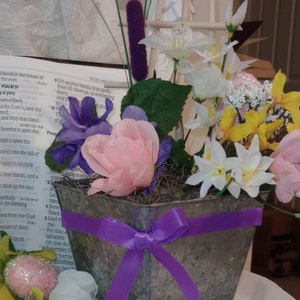 May include: A metal planter with a purple ribbon bow, filled with a variety of artificial flowers, including white, pink, yellow, and purple. The planter is sitting on a white surface with a book of Psalms visible in the background.