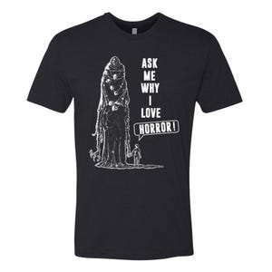 May include: Black t-shirt with a white graphic of a robed figure and a small person. The text "ASK ME WHY I LOVE HORROR!" is printed in white. The shirt is a crew neck style.