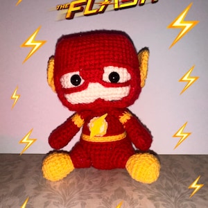 May include: A crocheted plush toy of The Flash, a superhero with a red and yellow costume. The toy has a big head with a red mask and a yellow lightning bolt on the chest. The toy is sitting on a brown surface with yellow lightning bolts around it. The text "THE FLASH" is written in yellow above the toy.