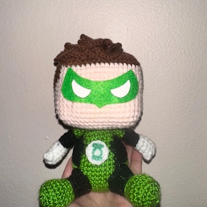 May include: Handmade crochet Green Lantern doll. The doll features a green and black costume, a green mask with white eye shapes, and brown hair. The Green Lantern symbol is on the chest. The doll is made of yarn.
