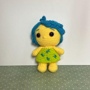 May include: A crocheted plush toy of a character with blue hair, yellow skin, and a green dress with blue stars. The toy has brown eyes and a small, pointed nose.