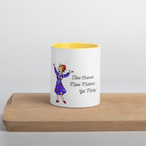 Può includere: Una tazza in ceramica bianca con un bordo giallo. La tazza presenta un'illustrazione di cartone animato di una donna con un vestito viola con accenti colorati. Il testo "Take Chances, Make Mistakes, Get Messy!" è stampato in nero sulla tazza.