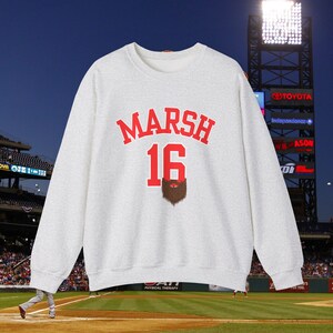 Puede incluir: Una sudadera gris claro con el nombre "MARSH" en letras rojas en bloque sobre el número "16" en rojo. Debajo del número hay un gráfico de barba marrón. El fondo muestra un estadio de béisbol por la noche.