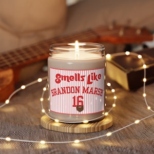 Puede incluir: Una vela encendida en un frasco de vidrio transparente con una etiqueta que dice "Smells Like Brandon Marsh 16". La etiqueta tiene un fondo a rayas rojas y blancas. La vela está sobre un pequeño posavasos de madera. Una guitarra y un libro están en el fondo.