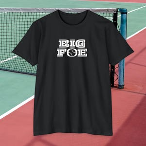 Puede incluir: Una camiseta negra de manga corta con el texto blanco BIG FOE impreso en el pecho. La letra O de la palabra FOE está sustituida por un gráfico blanco de una pelota de tenis con costuras visibles. La camiseta tiene cuello redondo y bajo recto.