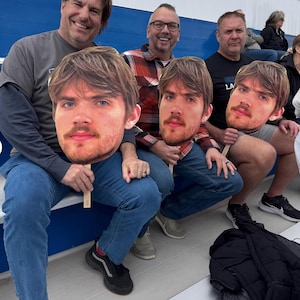 May include: Several people are seated, holding large cutouts of a man's face. The cutouts are attached to wooden sticks. The people are wearing casual clothing, including jeans and shirts. The background shows a blue and white bleacher setting.