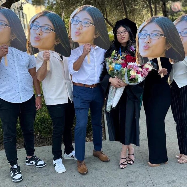 Pu&ograve; includere: Un gruppo di persone posa per una foto, alcune con grandi sagome di cartone di un volto. La persona al centro indossa una toga di laurea e tiene dei mazzi di fiori. Gli altri sono vestiti in abiti casual.
