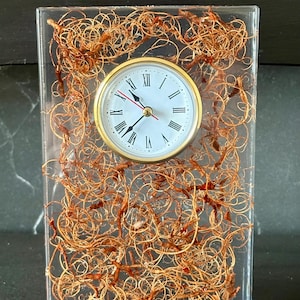 May include: A rectangular clear resin desk clock featuring a white face with black Roman numerals and gold-tone hands. The interior of the resin block is filled with a dense, tangled arrangement of thin, copper-colored metallic wire.