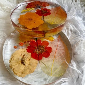 May include: Two clear resin coasters with dried flowers and leaves. One is a cup holder, and the other is a flat coaster. The flowers are orange, red, and yellow, and the leaves are a golden colour.