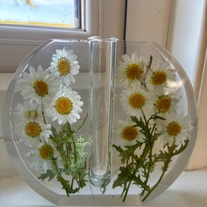 May include: A clear, oval-shaped vase with a central glass tube, showcasing white daisies with yellow centers encased in resin. The vase is filled with the flowers and green foliage, creating a decorative floral display.