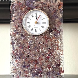 May include: A rectangular clear resin clock featuring embedded dried flower petals in shades of purple, pink, and white. The clock face is circular with a silver rim, white background, black Roman numerals, and the word QUARTZ printed in black.