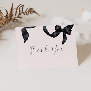 May include: A light pink card with a black bow illustration and the words "Thank You" in elegant script. The bow is at the top of the card. Dried foliage and a spool of ribbon are in the background.