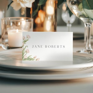 Könnte beinhalten: Eine weiße Tischkarte mit dem Namen "JANE ROBERTS" in Schwarz gedruckt. Die Karte ist mit aquarellfarbenen rosa Blumen und grünen Blättern verziert. Die Karte liegt auf einem weißen Teller, mit Kerzen und Gläsern im Hintergrund.