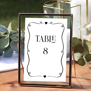 May include: A framed table number card with the word "TABLE" and the number "8" in black font. The card is white with a decorative black border and heart accents. The frame is gold-colored.