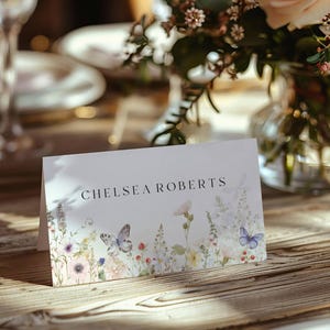Könnte beinhalten: Eine weiße Tischkarte mit dem Namen "CHELSEA ROBERTS" in Schwarz gedruckt. Die Karte zeigt ein Aquarell-Blumenmuster mit Schmetterlingen in Blau-, Rosa- und Gelbtönen. Die Karte liegt auf einem Holztisch, mit einem Blumengesteck im Hintergrund.