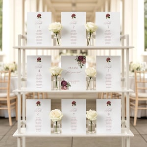 May include: A white three-tiered wooden display stand holding eight rectangular table seating cards and one central sign. Each card features a dark red floral illustration, table numbers one through eight, and guest names. Small glass vases with white roses are placed between the cards.
