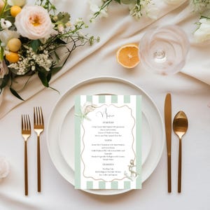 Pode incluir: Um cartão de menu com um fundo listrado em verde menta e branco, com um design de concha e âncora. O menu lista entradas, pratos principais e sobremesas. Talheres dourados estão dispostos em um prato branco, com flores e uma fatia de limão.