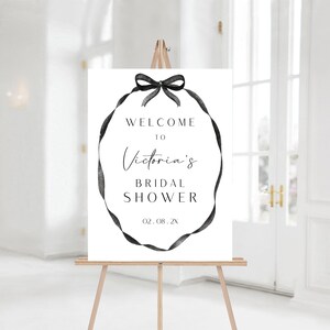 May include: White welcome sign with a black ribbon wreath and the text "Welcome to Victoria's Bridal Shower 02.08.2X"