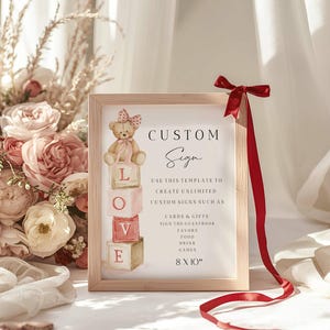 Peut inclure: Un panneau encadré avec le texte "CUSTOM Sign" et une illustration d'ours en peluche. Le panneau comprend le texte "LOVE" sur des blocs empilés et du texte supplémentaire sur la création de panneaux personnalisés. Un ruban rouge est attaché au cadre. Le panneau mesure 20 x 25 cm.