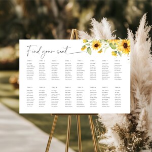 Pode incluir: Um quadro de assentos de casamento branco com as palavras "Find your seat" em escrita elegante. O quadro é decorado com girassóis amarelos e folhas verdes. O quadro é exibido em um cavalete dourado.