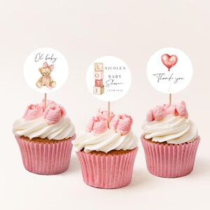 Peut inclure: Trois cupcakes avec des caissettes roses et un glaçage blanc, surmontés de décorations florales roses. Chaque cupcake a un topper circulaire avec du texte et des illustrations : un ours en peluche, le texte "NICOLAS BABY SHOWER 14 FEB 2023", et un ballon cœur.