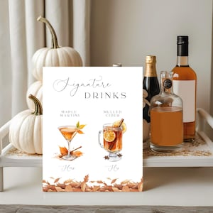 以下が含まれることがあります： 「Signature Drinks」の文字と、メープルマティーニとホットサイダーのイラストが描かれた白い看板。看板は秋の葉で飾られ、白いカボチャとアルコールボトルの前に置かれています。