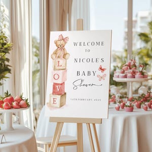 Peut inclure: Panneau de bienvenue pour une baby shower sur un chevalet. Le panneau présente un ours en peluche, le mot "LOVE" sur des blocs et le texte "Bienvenue à la baby shower de Nicole" avec la date.