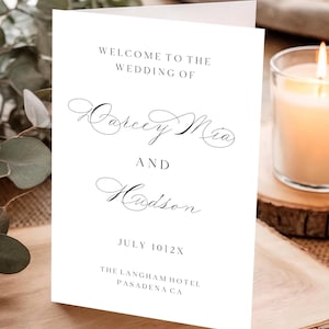 Pode incluir: Um convite de casamento branco com letras pretas. O texto diz: "Bem-vindos ao casamento de Darcey Mia e Hudson, 10/2X de julho, The Langham Hotel, Pasadena CA". Uma vela acesa e vegetação ao fundo.