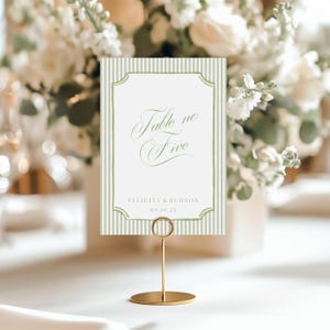 May include: A rectangular table number card featuring a white background with vertical sage green stripes and a sage green border. The text reads Table no Five, Felicity & Hudson, and 09.19.2X. The card is held by a thin gold metal stand.