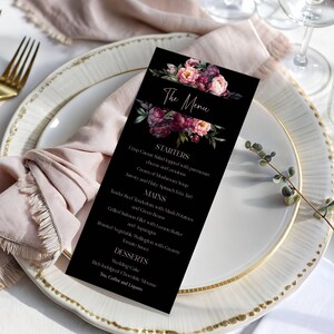 May include: A black menu card with floral accents, featuring the text "The Menu" and a list of starters, mains, and desserts. The card is placed on a white plate with a gold rim, alongside a pink napkin and gold forks.