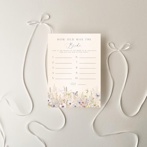 May include: A cream-colored bridal shower game card with the text "How Old Was The Bride". The card features a floral design at the bottom and lines for guests to write their guesses. White ribbons are arranged around the card.