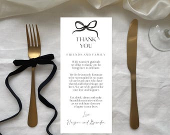 May include: A gold-colored fork with a black ribbon tied around it, and a gold-colored knife are displayed next to a white card with the words "Thank You" and a message. The items are arranged on a white cloth.