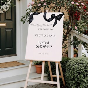 Pode incluir: Uma placa de boas-vindas para um chá de panela, exibida em um cavalete de madeira. A placa é rosa claro com texto preto e um design de fita preta. O texto diz "She's tying the knot" e "Welcome to Victoria's Bridal Shower 7th Feb 202X".