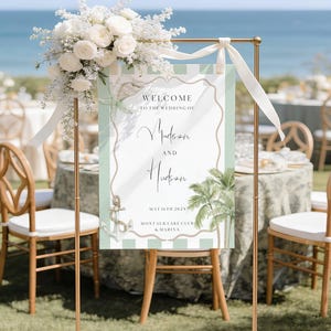 May include: A gold wedding sign with a white and pale green striped design. The sign displays the text "Welcome to the Wedding of Madison and Hudson, May 16th 202X, Montauk Lake Club & Marina." A white floral arrangement and ribbon are at the top.