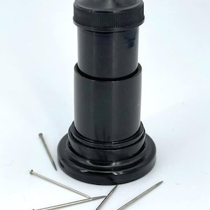 Peut inclure: Un outil de couture cylindrique noir avec un dessus conique et une large base. Plusieurs épingles argentées à petites têtes rondes sont éparpillées autour de la base de l'outil. L'outil semble être en plastique.
