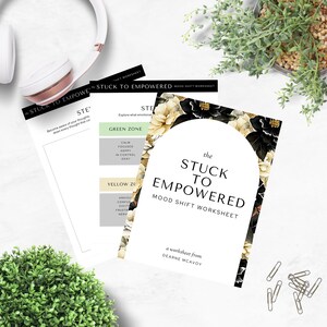 May include: A stack of white worksheets titled "The Stuck to Empowered Mood Shift Worksheet" with floral accents. The worksheets are accompanied by a pair of white headphones, greenery, and paper clips.