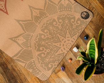 May include: A cork yoga mat with a brown mandala design and the text "Yogi Rio" printed on the top right corner. The mat is lying on a wooden surface with a potted plant and crystals.
