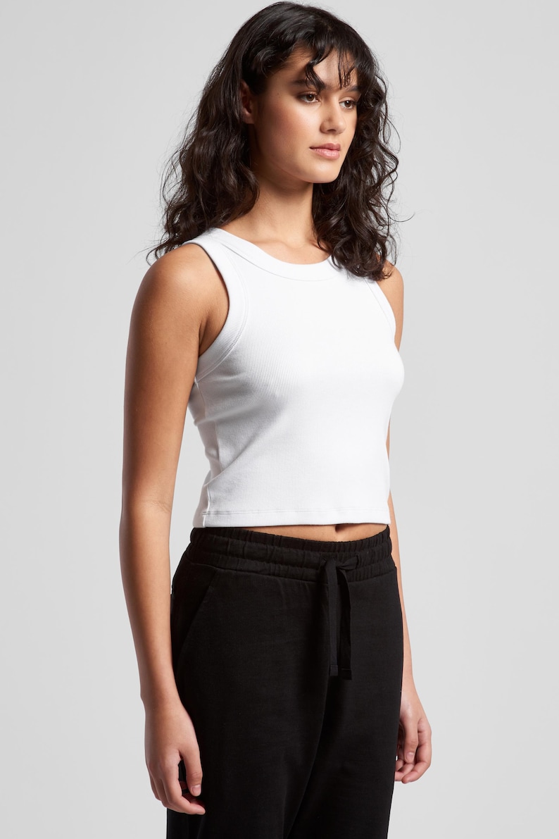 May include: A white ribbed tank top with a crew neck and a cropped fit. The model is also wearing black sweatpants. The top is a solid color and appears to be made of a soft, stretchy material.